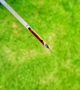 A close-up photograph of a dipstick with a drop of black oil on the end, indicating that the oil needs to be changed. The dipstick is held in front of a blurred background of green grass.