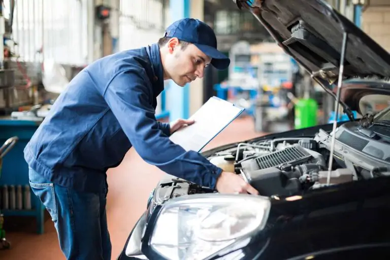 A mechanic in a blue uniform inspects the engine of a black car, clipboard in hand. He is checking the car's fluids. 