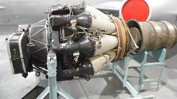 A close-up of a large black and white jet engine, with a red circle in the background. It is sitting on a stand. The engine is made of metal and has a lot of wires and tubes attached to it. It is a very powerful engine and looks like it could power a large aircraft.