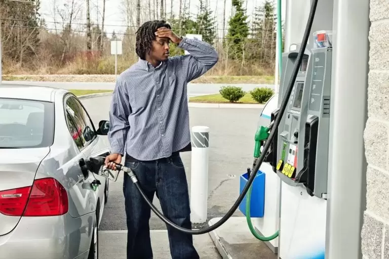 A man at a gas station holds a fuel pump to his car while looking at the camera with a frustrated expression, a hand on his head. 