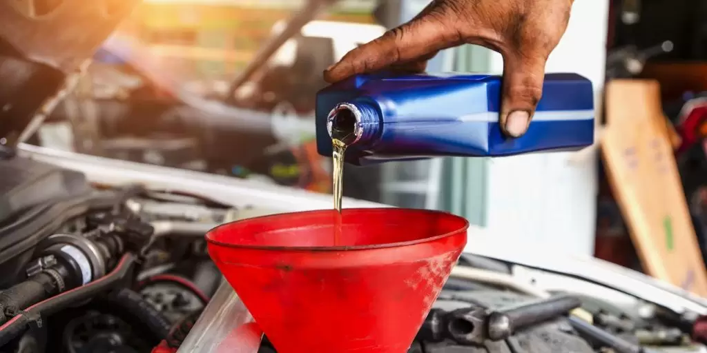 A mechanic is pouring oil into a car engine. The oil is amber-colored and the engine is dark and dirty. The mechanic is wearing a blue shirt and his hand is holding the oil bottle. The background is blurry and there is a bright light coming from the left side of the image.