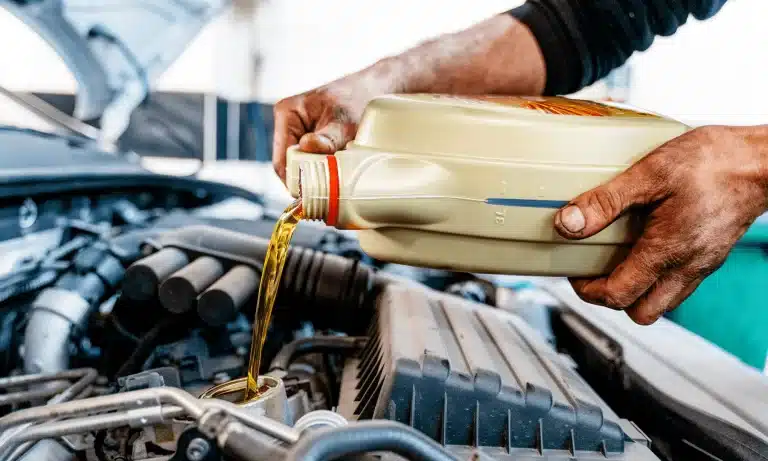 A mechanic wearing black is pouring oil into a car engine, close-up. The mechanic's hands are dirty and the oil is amber-colored. The engine bay is dark and the light is coming from the left side of the image.